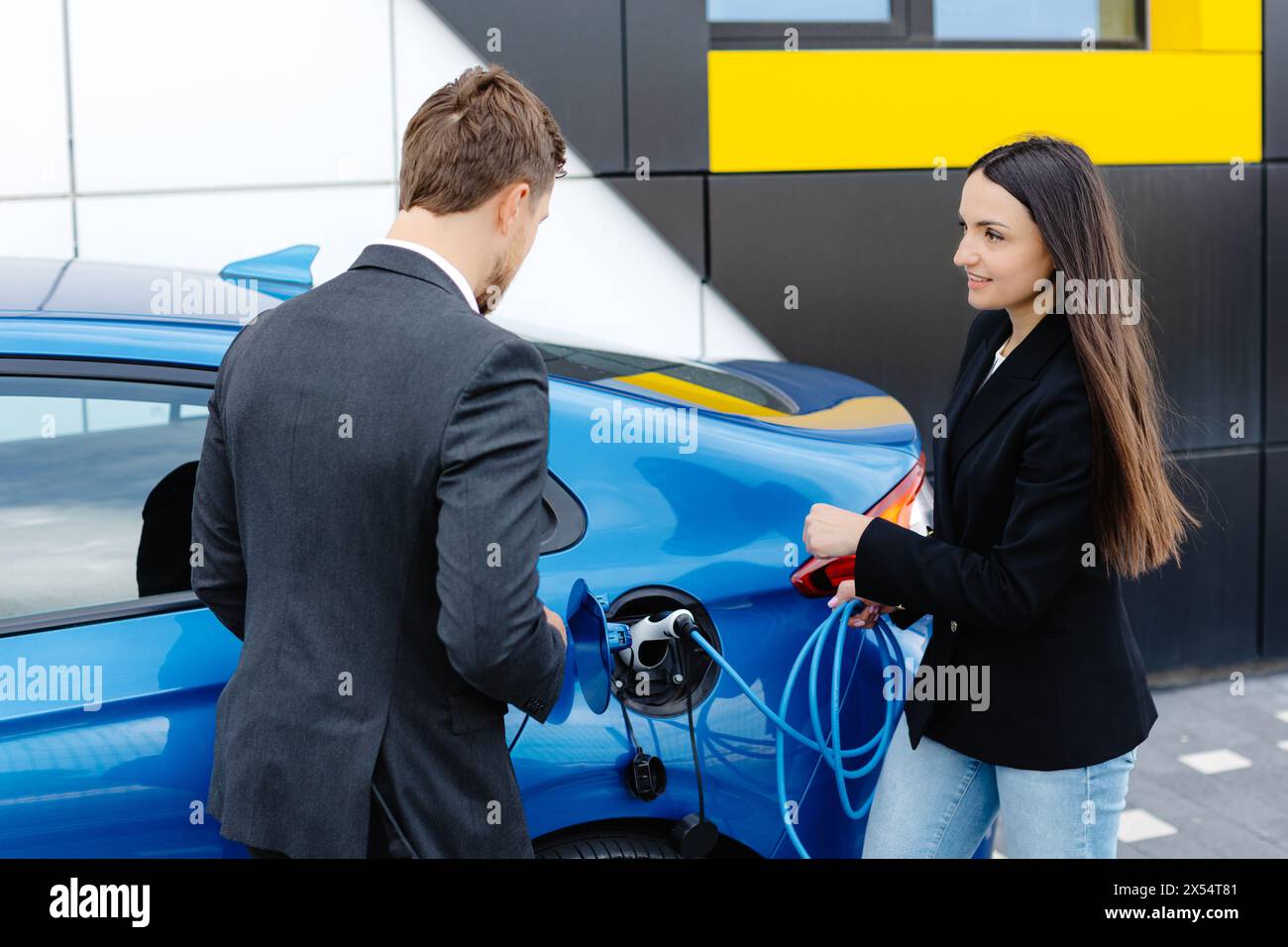 Responsabile vendite che mostra come caricare l'auto elettrica a una giovane donna cliente, collegando il filo alla presa per auto presso la concessionaria auto all'aperto. Foto Stock