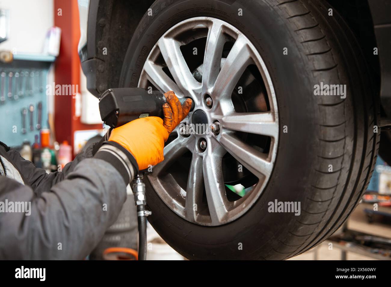 Anonimo meccanico maschile in guanti e in giacca, mentre si trova vicino all'auto sollevata in garage e svitando la ruota dell'auto con l'attrezzo in mano Foto Stock