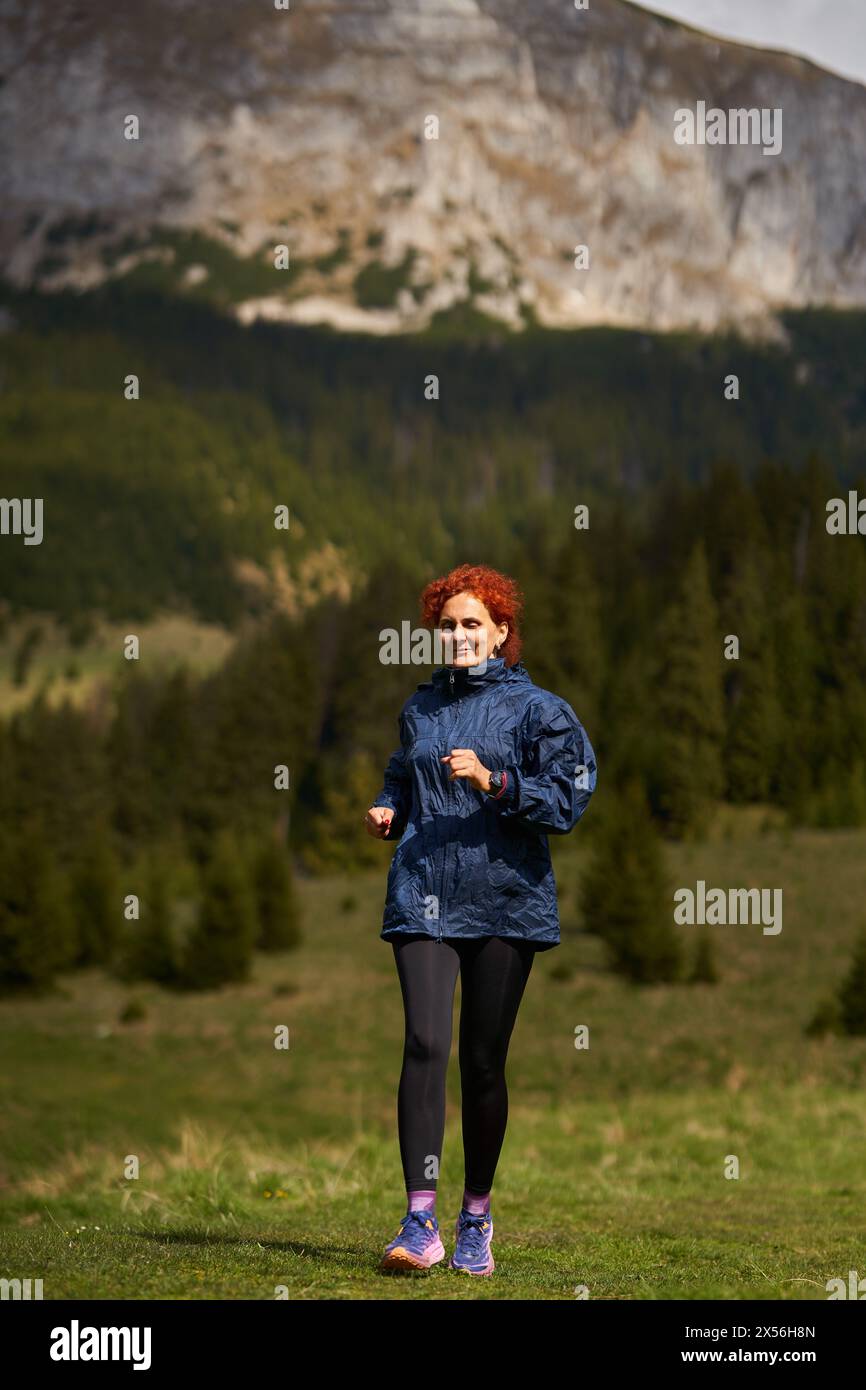 Donna trail runner su un percorso di montagna che corre su un prato con foreste e montagne alle spalle Foto Stock