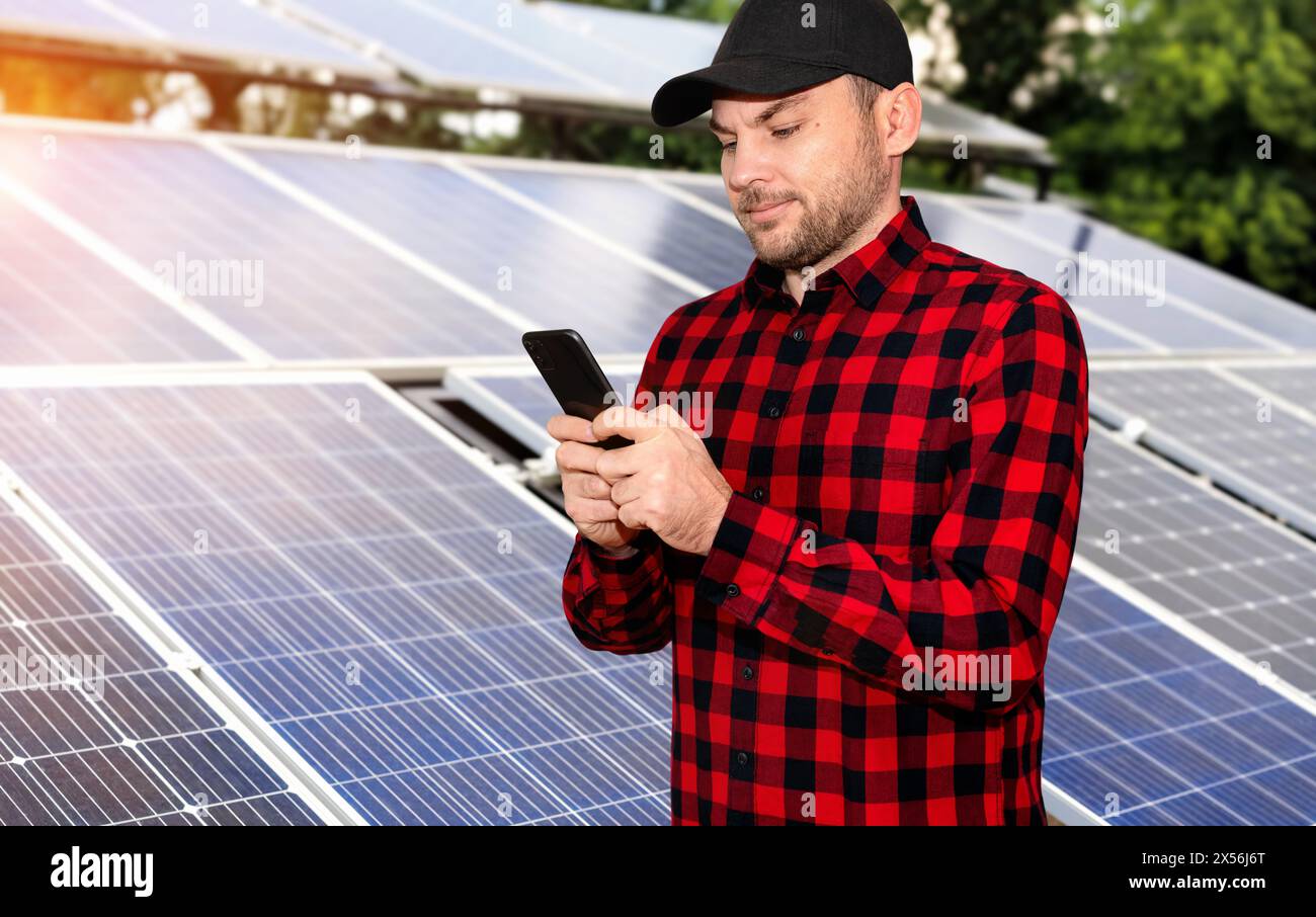 Uomo che indossa una camicia casual rossa-nera e un berretto nero usando il suo smartphone accanto ai pannelli a energia solare nella centrale solare. Foto Stock