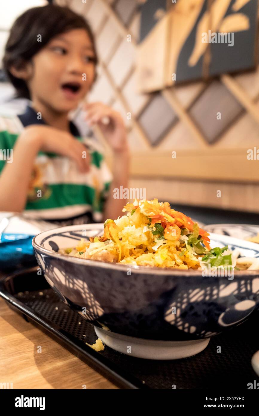 Una ciotola di menu' di noodles servito al ristorante Giapponese, con un bambino felice del Sud-Est Asiatico sullo sfondo Foto Stock