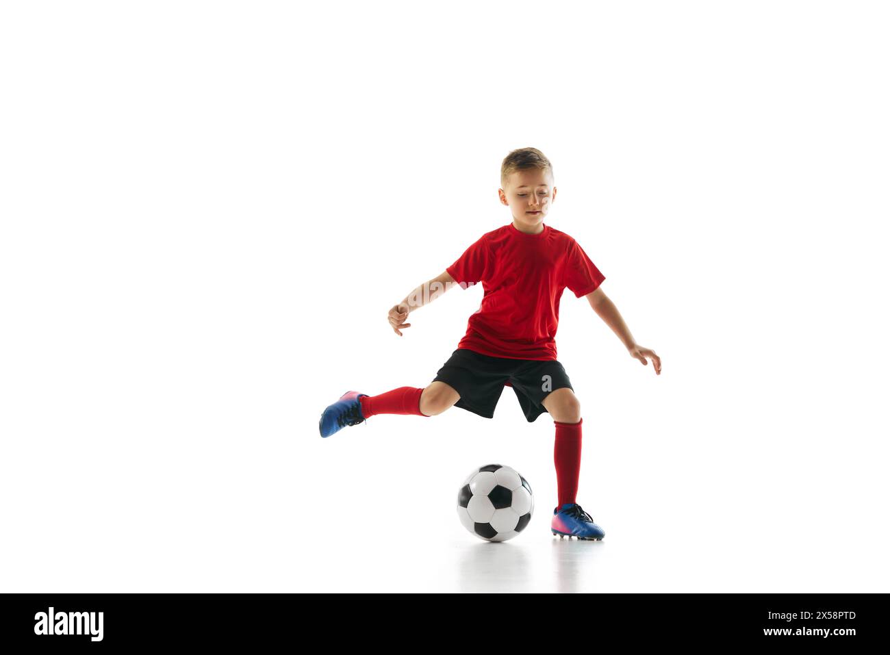 Il colpo dinamico di un bambino in uniforme sportiva gioca a calcio, allenando la palla passante su sfondo bianco. Foto Stock