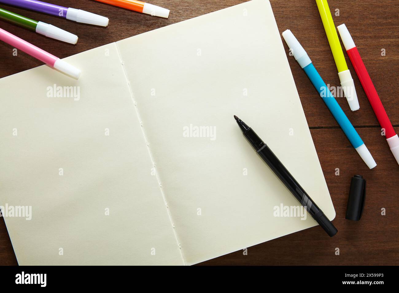 un bozzetto vuoto aperto con un pennarello nero pronto all'uso e una serie di pennarelli colorati sul tavolo di legno Foto Stock