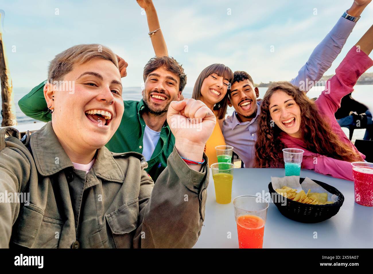 Selfie per gruppi di amici diversi - ridendo e gustando un drink in spiaggia, mostrando gioia e solidarietà in un ambiente informale all'aperto. Foto Stock