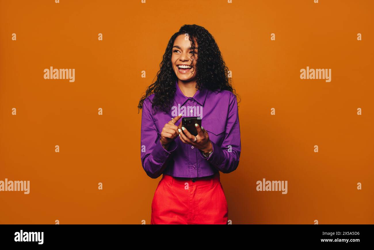 Una donna allegra in un abbigliamento casual si trova in uno studio su uno sfondo arancione brillante. Utilizza il suo moderno smartphone per navigare sui social media. Foto Stock