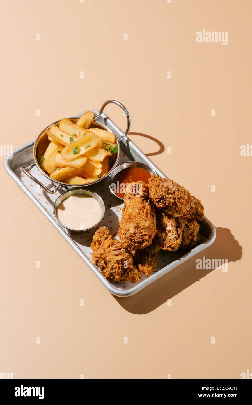 Un vassoio contenente ali di pollo fritte dorate abbinate a patatine fritte condite, con salse cremose e piccanti presentate su sfondo beige Foto Stock