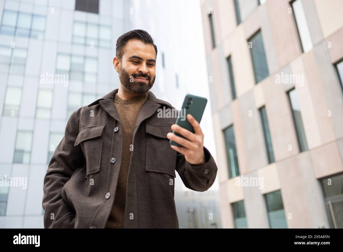 Un uomo sorridente in un cappotto elegante interagisce con il suo smartphone in una vivace strada della città, con edifici moderni sullo sfondo Foto Stock