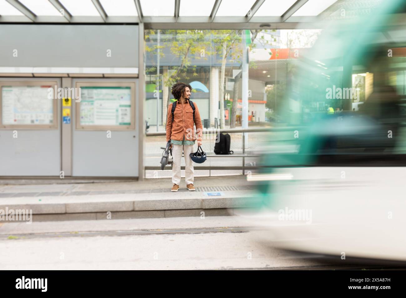 Un uomo si trova presso una fermata dell'autobus con in mano uno scooter elettrico e un casco, con il movimento sfocato di un autobus che passa, suggerendo la mobilità urbana Foto Stock