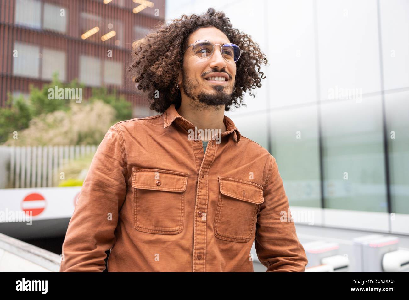 Un giovane uomo con capelli ricci e occhiali sguardi attentamente, vestito con un'elegante camicia marrone su uno sfondo urbano Foto Stock