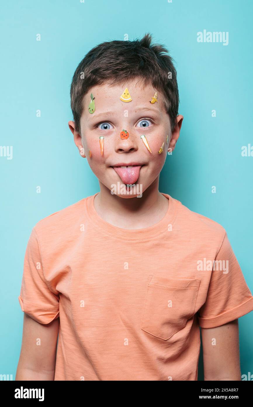 Un ragazzo giocoso crea un volto divertente con la lingua fuori e gli adesivi di frutta sulla pelle su uno sfondo azzurro Foto Stock