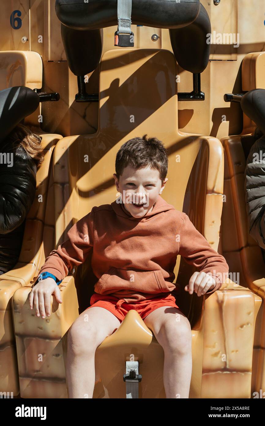 Un ragazzo allegro che indossa una felpa con cappuccio e pantaloncini corti sorride mentre è seduto su un parco divertimenti dorato durante una calda giornata estiva. Foto Stock