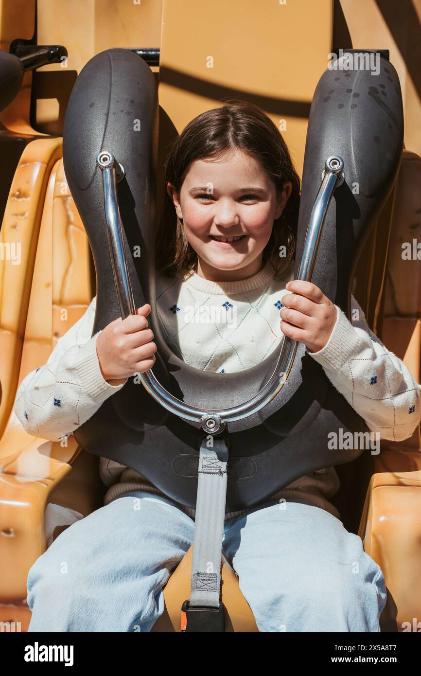 Una giovane ragazza allegra con un sorriso luminoso è al sicuro nel suo posto in un parco divertimenti, pronta a vivere un'esperienza emozionante durante un'estate soleggiata Foto Stock