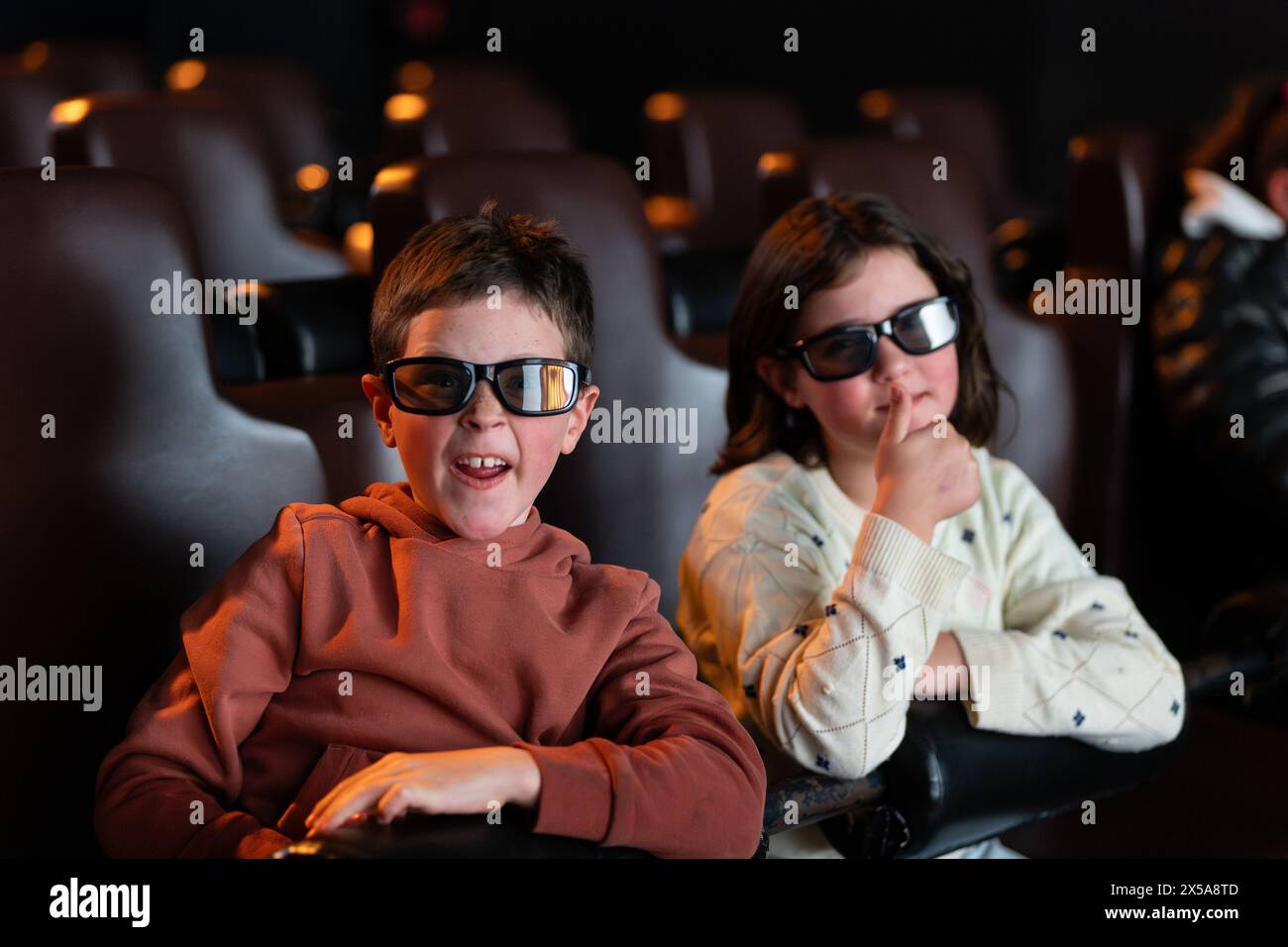 Due bambini, un ragazzo e una ragazza, indossano occhiali 3D e guardano un film con espressioni intriganti durante una giornata estiva al cinema. Foto Stock