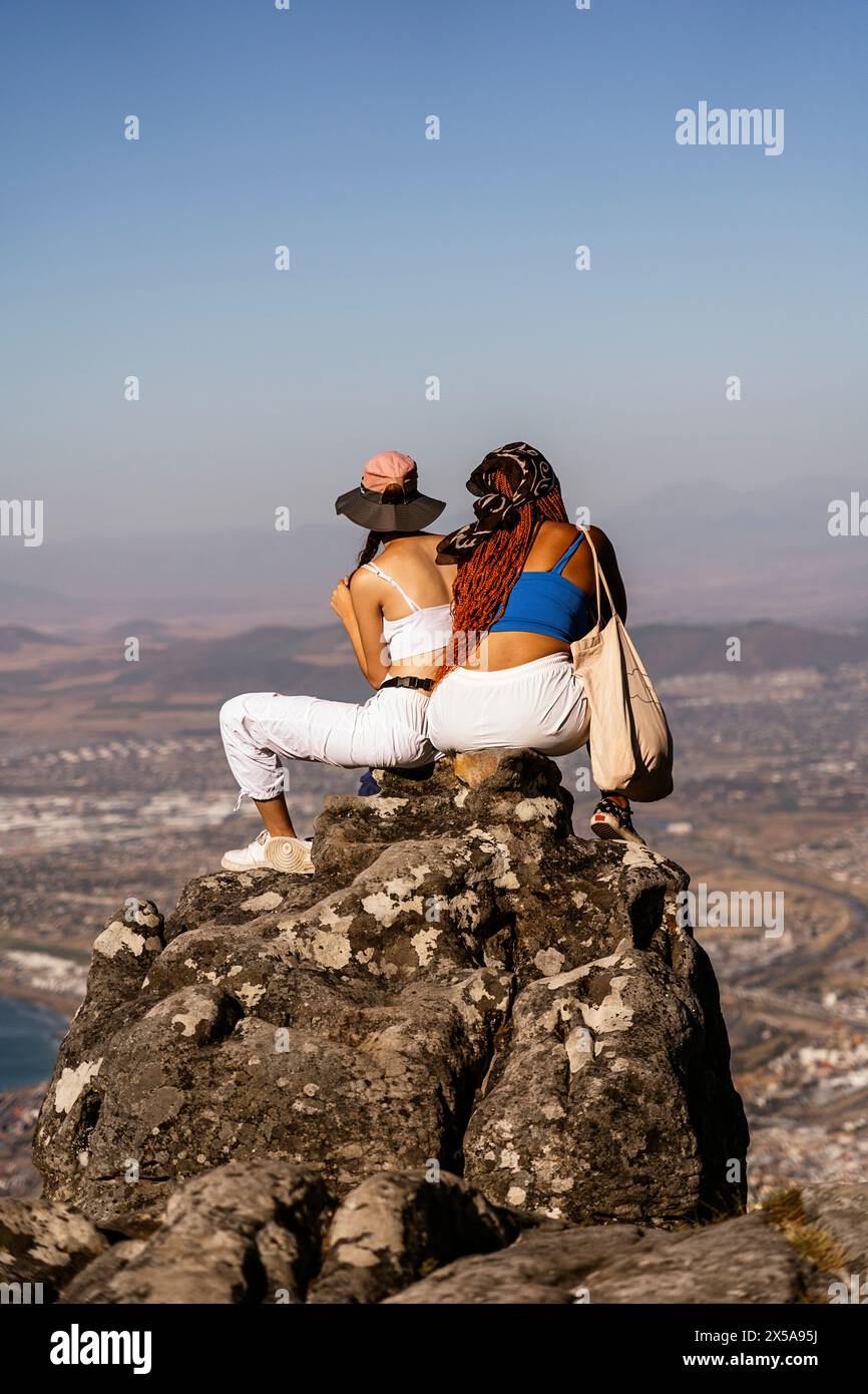 Due persone si siedono in cima a una montagna, osservando un vasto paesaggio sottostante. Immersi serenamente nella vista, sono retroilluminati da un cielo limpido. Foto Stock