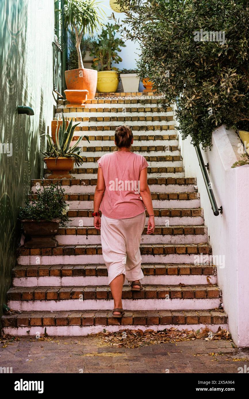 Vista posteriore di una donna irriconoscibile che sale le scale di mattoni circondate da piante in vaso in un vicolo in stile mediterraneo, guardando lontano dalla macchina fotografica. Foto Stock