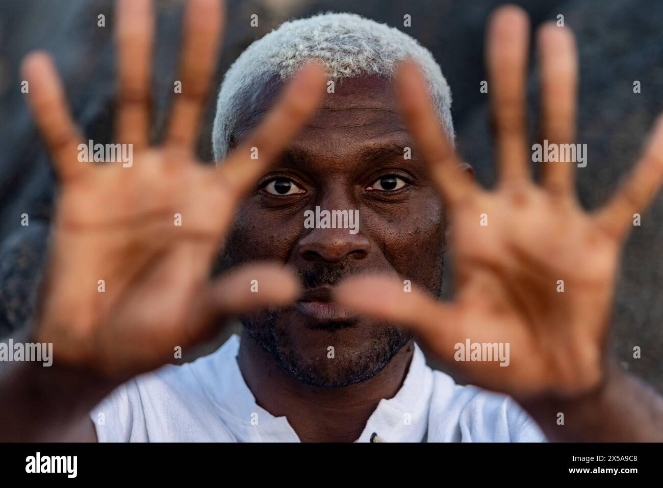 Un avvincente ritratto ravvicinato di un uomo afroamericano con i suoi sorprendenti capelli bianchi, che guarda attraverso le sue dita allungate Foto Stock