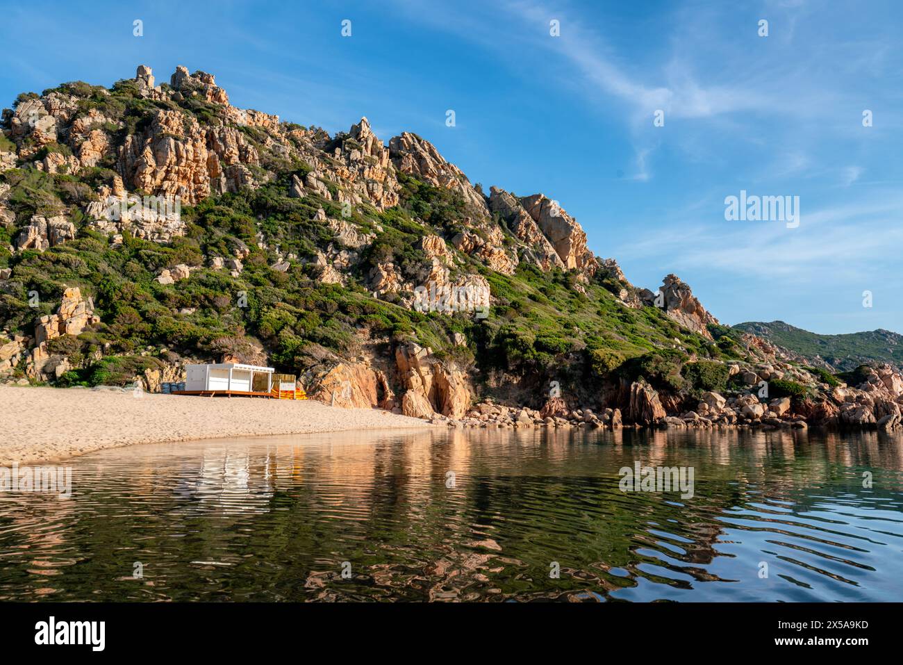 Tranquilla spiaggia li Cossi nella Costa Paradiso, in Sardegna, con aspre colline e un cielo azzurro che si riflette sulle acque calme Foto Stock