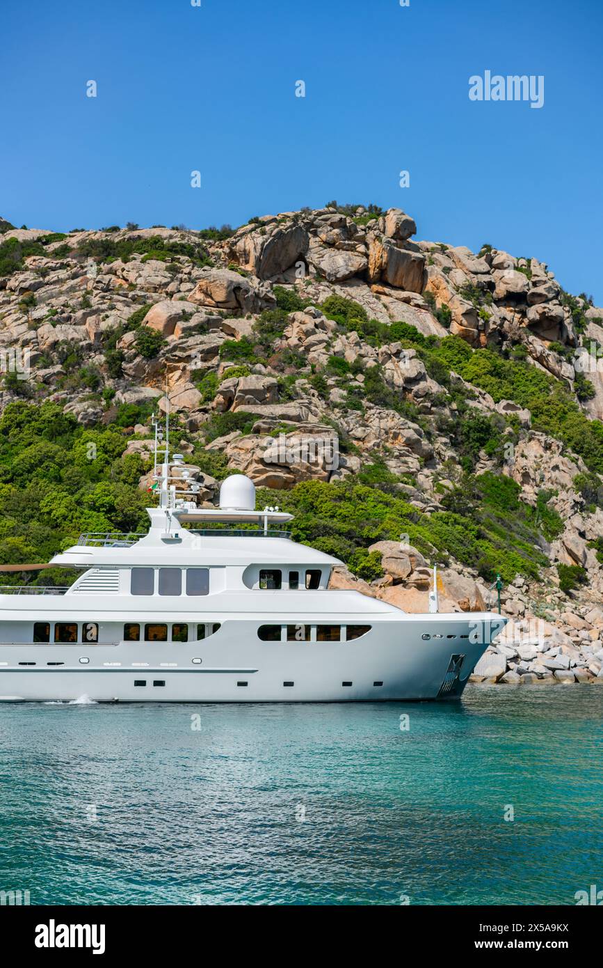 Un elegante yacht bianco di lusso galleggia sulle limpide acque turchesi della Sardegna, Italia, con maestose colline rocciose sullo sfondo, grande Pevero Beac Foto Stock