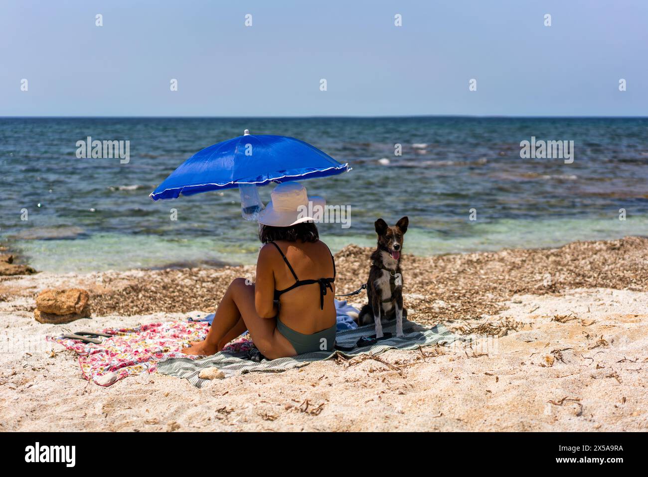 Una donna si rilassa sotto un ombrello blu con il suo fedele cane su una spiaggia soleggiata in Sardegna, Italia, incarnando lo spirito del tempo libero estivo Foto Stock