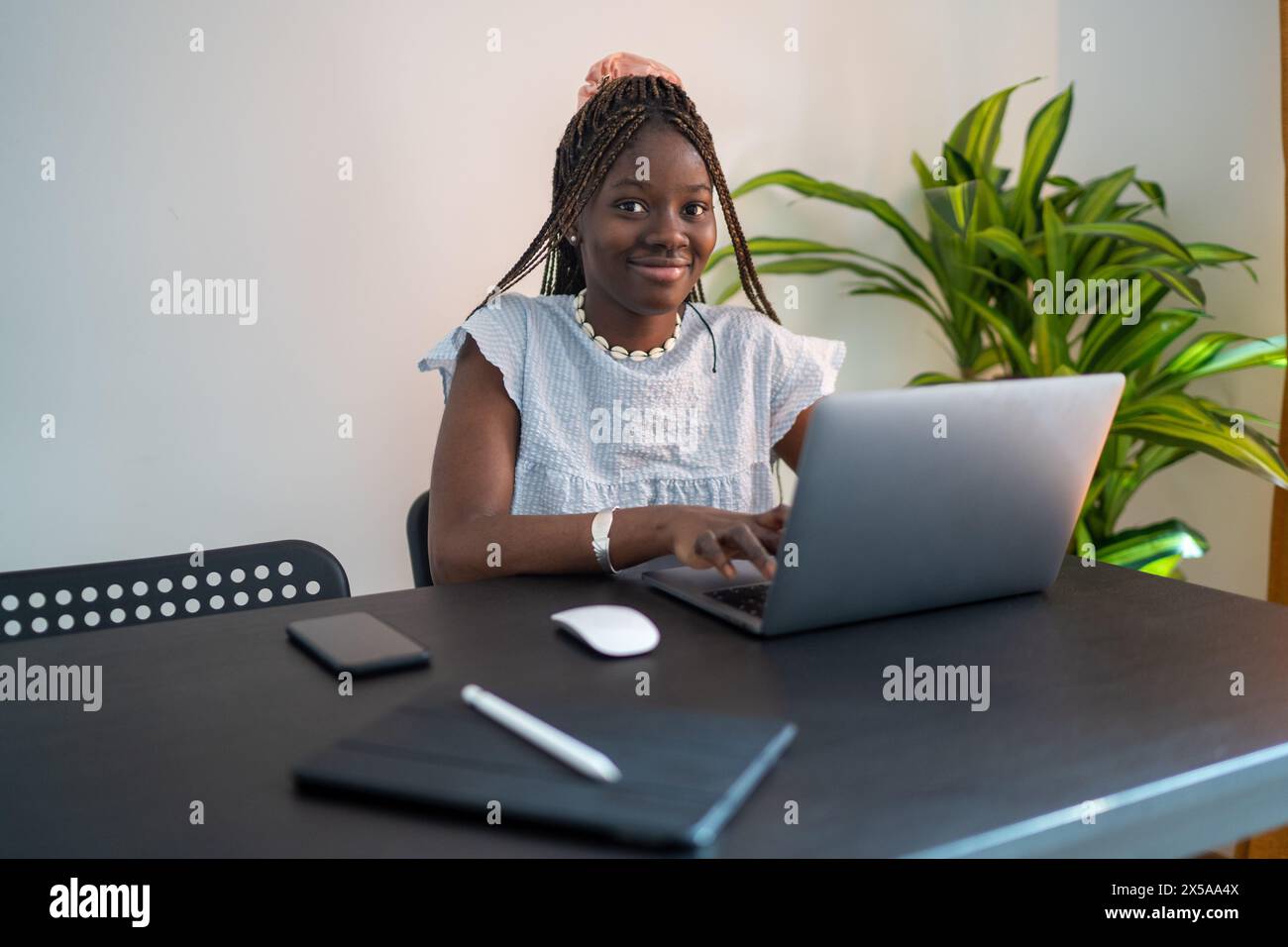 Una donna nera sorridente con i capelli intrecciati lavora con il suo laptop in un ufficio domestico, trasudando produttività e comfort Foto Stock