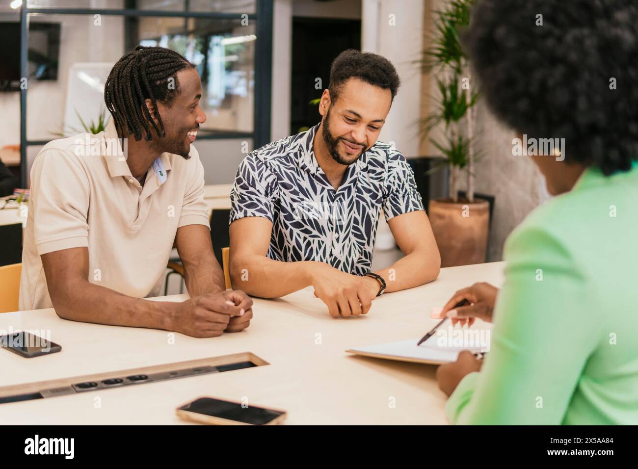 Due uomini sorridenti che si impegnano con un collega in un ambiente di coworking moderno e pieno di piante Foto Stock