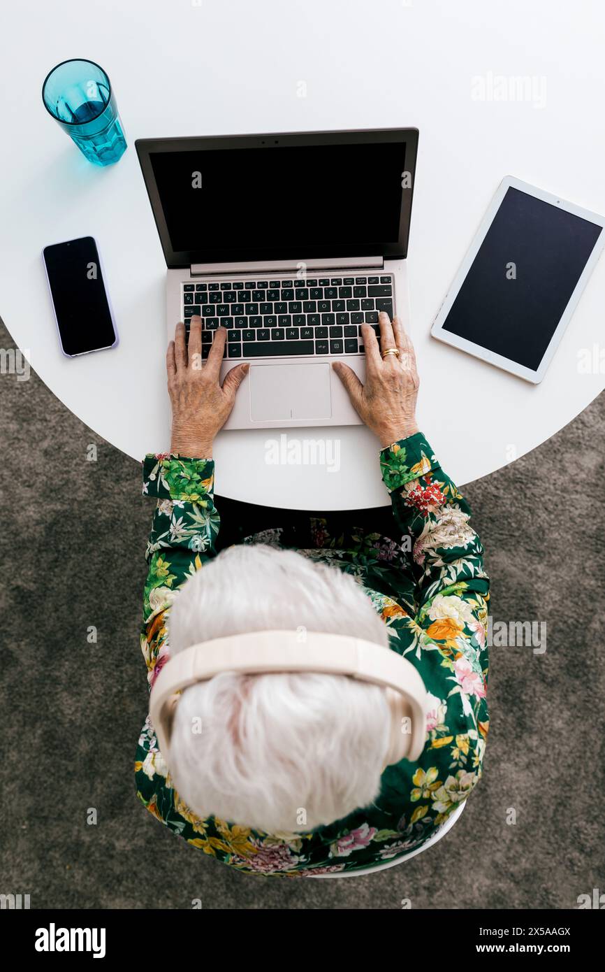 Una donna anziana con una camicia floreale viene vista dall'alto mentre lavora sul suo computer portatile a casa, con telefono e tablet nelle vicinanze Foto Stock