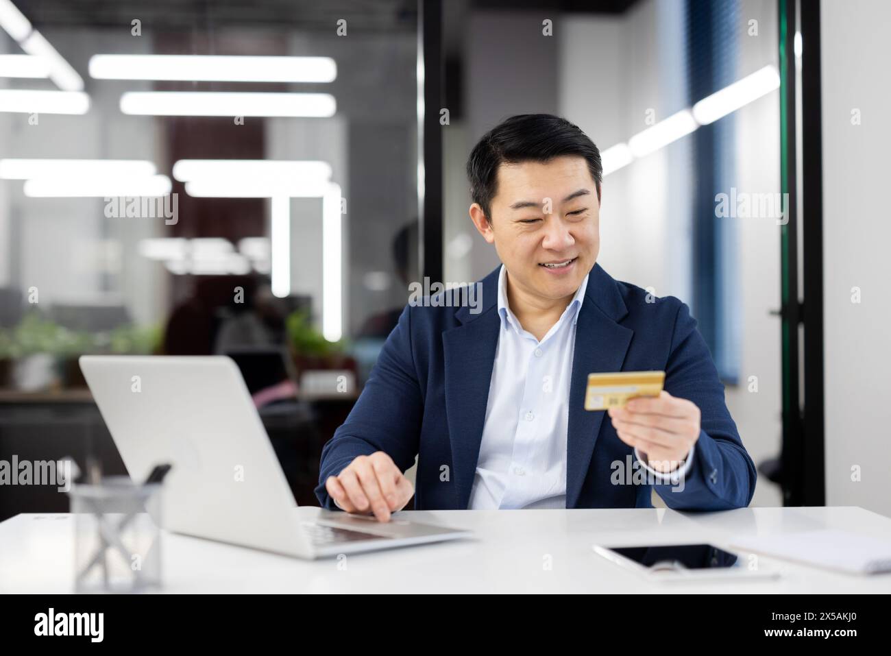 Uomo d'affari asiatico sorridente in un ambiente moderno che utilizza una carta di credito per gli acquisti online. Sembra contento mentre si impegna in una transazione di e-commerce. Foto Stock