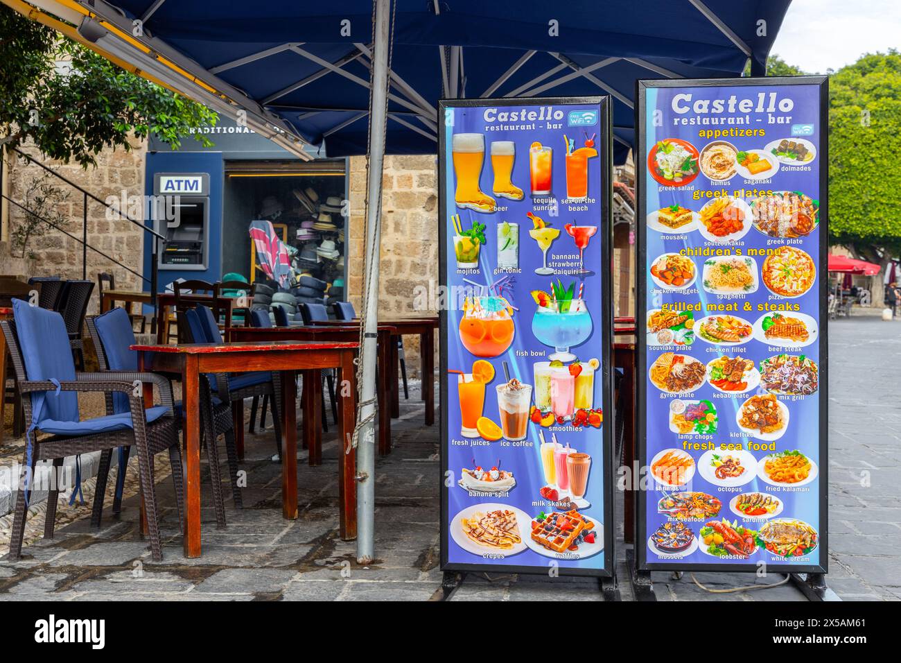 Rodi, Grecia - 12 giugno 2023: Cartelli del menu per cibo e bevande presso il ristorante chiuso con sedie e tavoli. Bancomat in background Foto Stock