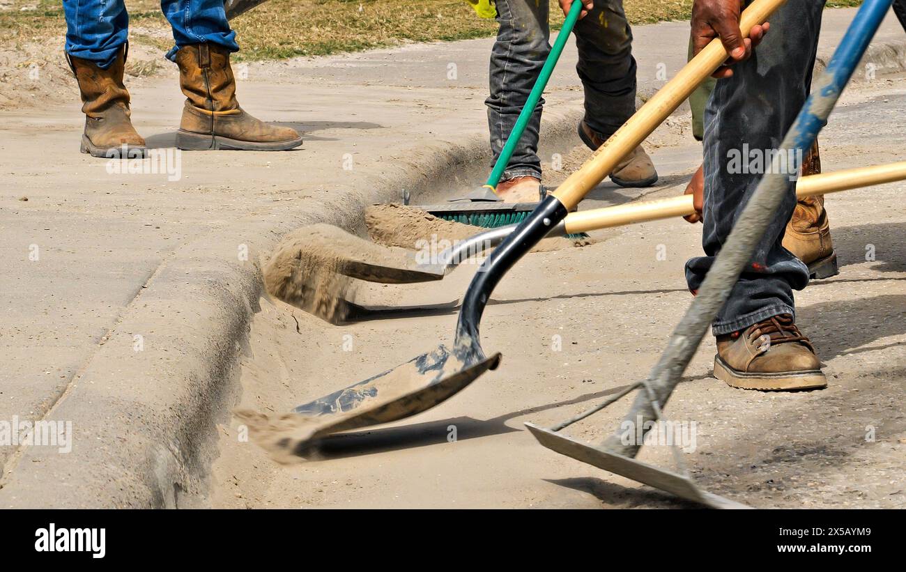 Gli operai edili utilizzano pale e scopa per rimuovere lo sporco dalla strada lungo il marciapiede di un cantiere. Primo piano di stivali, pale, sporcizia e marciapiede. Foto Stock