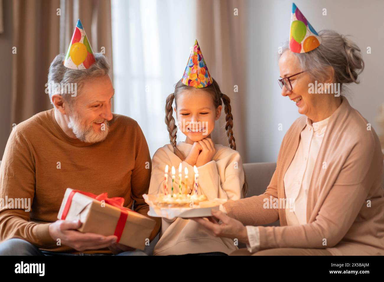 Nonni gioiosi che celebrano il compleanno delle nipoti con torta e regalo al chiuso Foto Stock