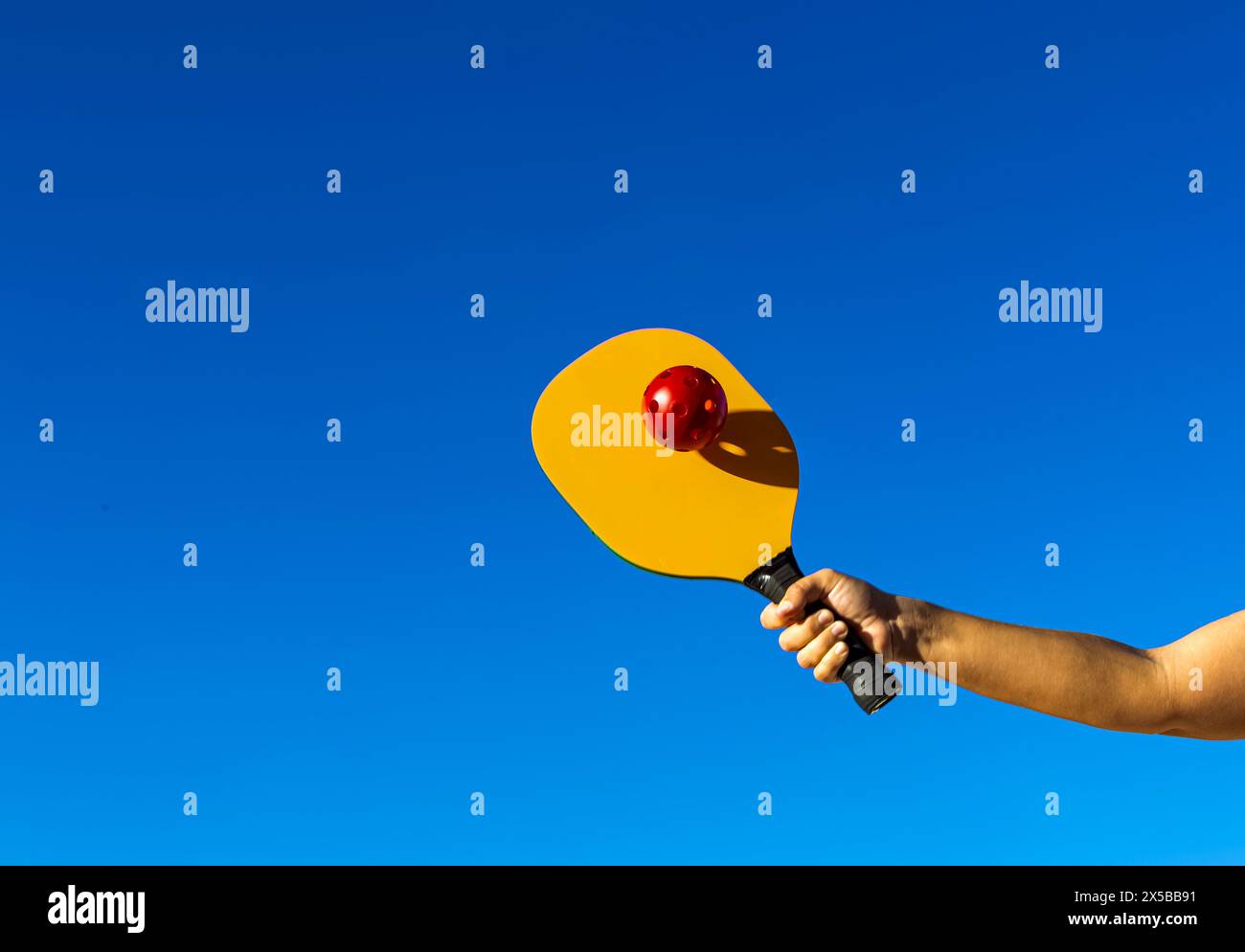 Braccio che regge il picchetto e colpisce la palla. Racchetta da picchetto gialla che colpisce una palla rossa, con il cielo di sfondo e lo spazio di copia Foto Stock