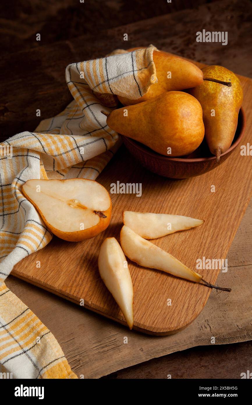 Tagliere in legno con fette e metà di frutta di pera matura, ciotola di argilla con pere e asciugamano da cucina giallo su fondo di legno vintage. Foto Stock