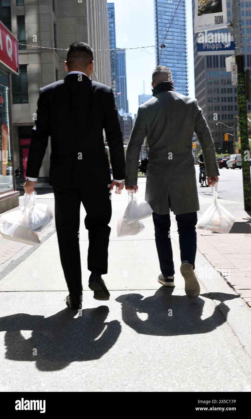 Due uomini in abiti da lavoro camminano lungo una strada della città portando borse di plastica con piatti per il pranzo all'interno Foto Stock