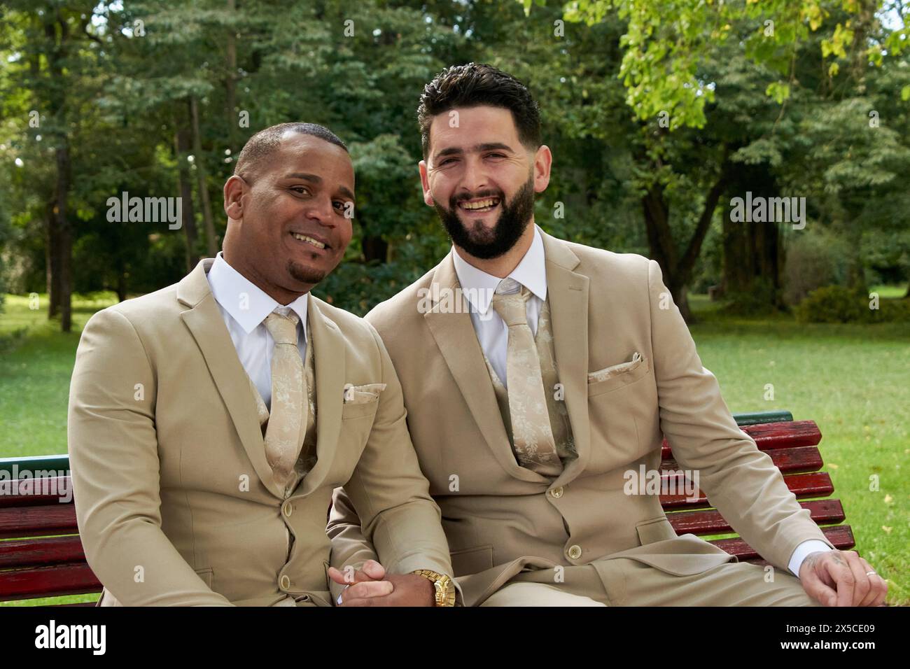 Ritratto di una coppia appena sposata della comunità gay, entrambi vestiti formali e sorridenti alla macchina fotografica, tenendosi per mano. Foto Stock