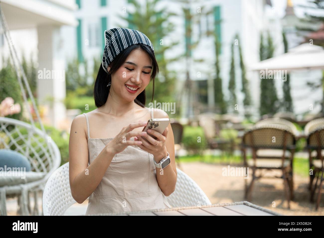 Una bella donna asiatica con un bel vestito si siede in un bellissimo giardino inglese usando il suo smartphone, chiacchierando con i suoi amici o leggendo messaggi Foto Stock