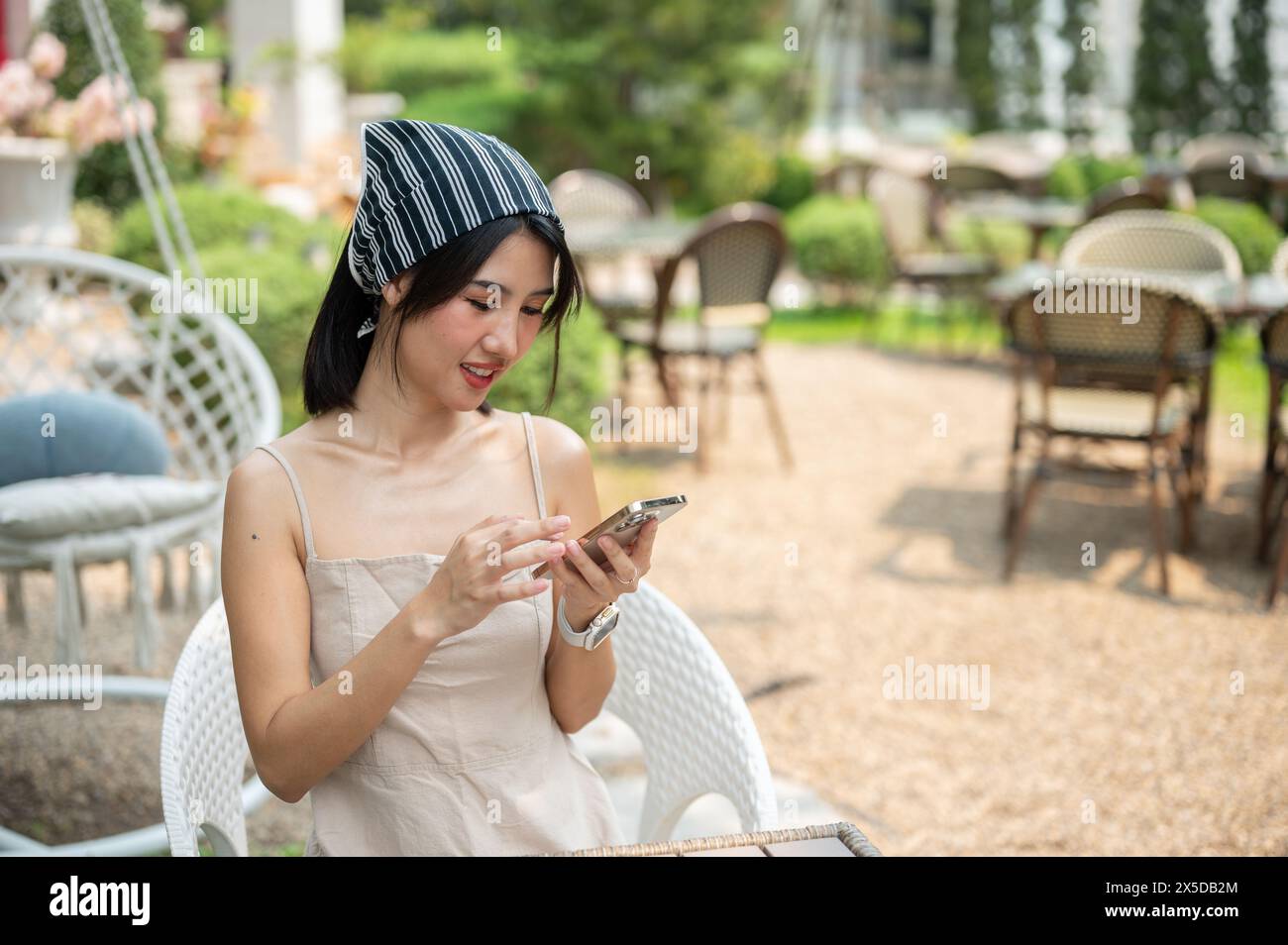 Una bella donna asiatica con un bel vestito si siede in un bellissimo giardino inglese usando il suo smartphone, chiacchierando con i suoi amici o leggendo messaggi Foto Stock