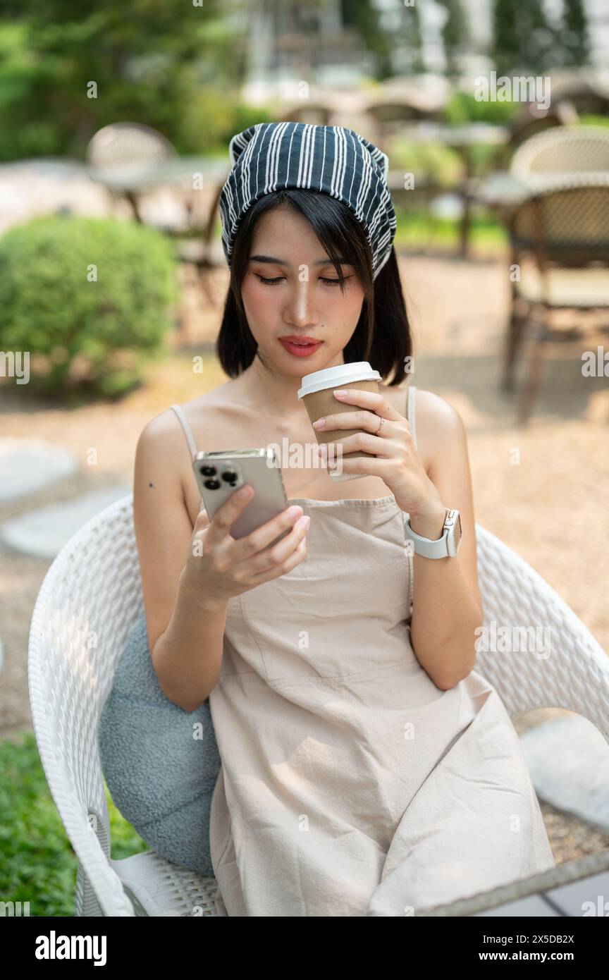 Una bella donna asiatica con un grazioso abito minimal si siede in un bellissimo giardino inglese sorseggiando un caffè e utilizzando il suo smartphone. chiacchierare, social media, mese Foto Stock