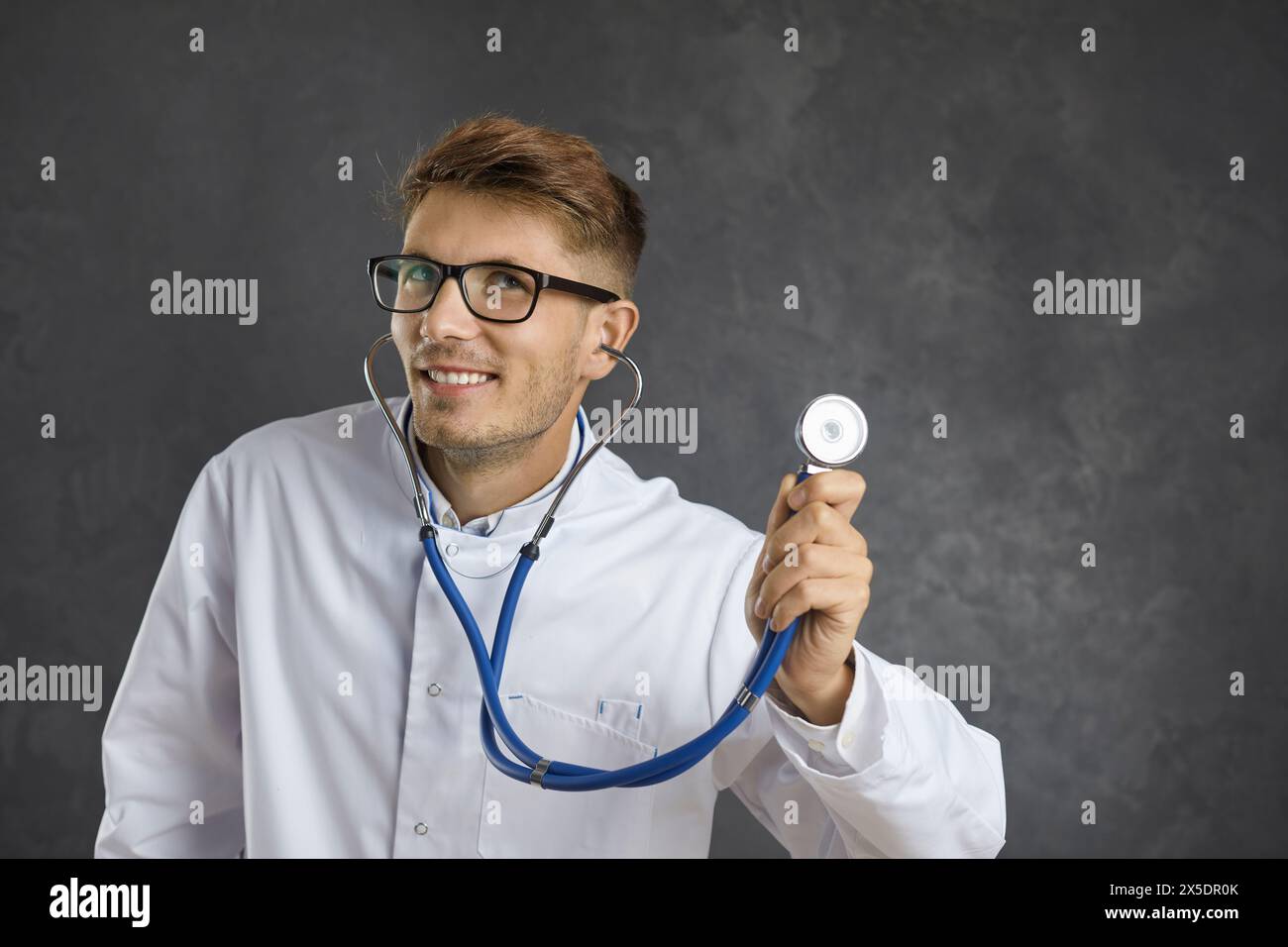Giovane medico maschio sorridente che ascolta attraverso lo stetoscopio e pensa a qualcosa di bello. Foto Stock