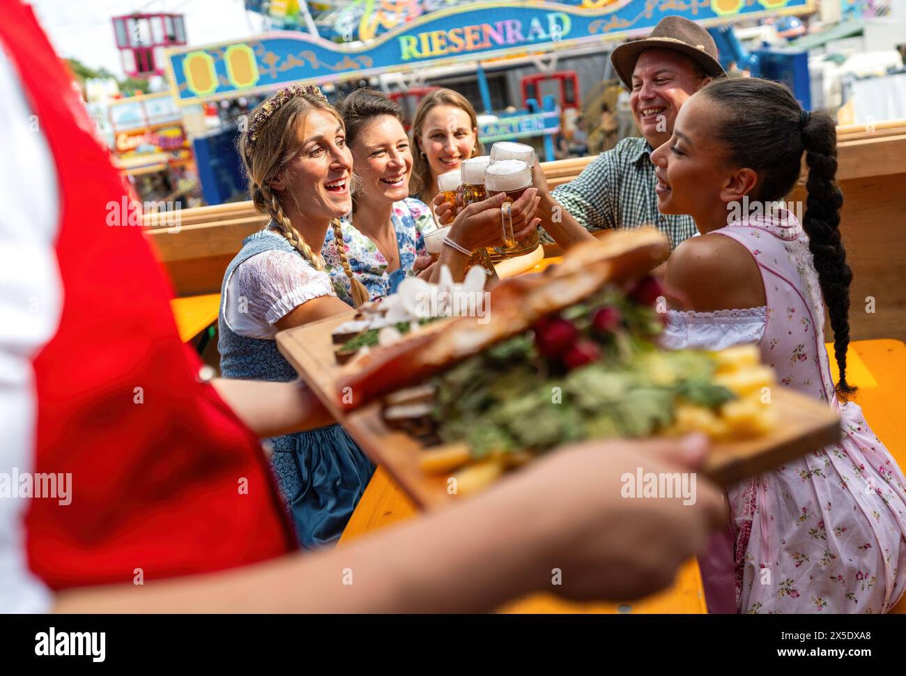 Amici al tavolo del festival oktoberfest che ricevono un vassoio di cibo tradizionale, brindano gioiosamente con birra, giostre di divertimento sullo sfondo a monaco di baviera Foto Stock