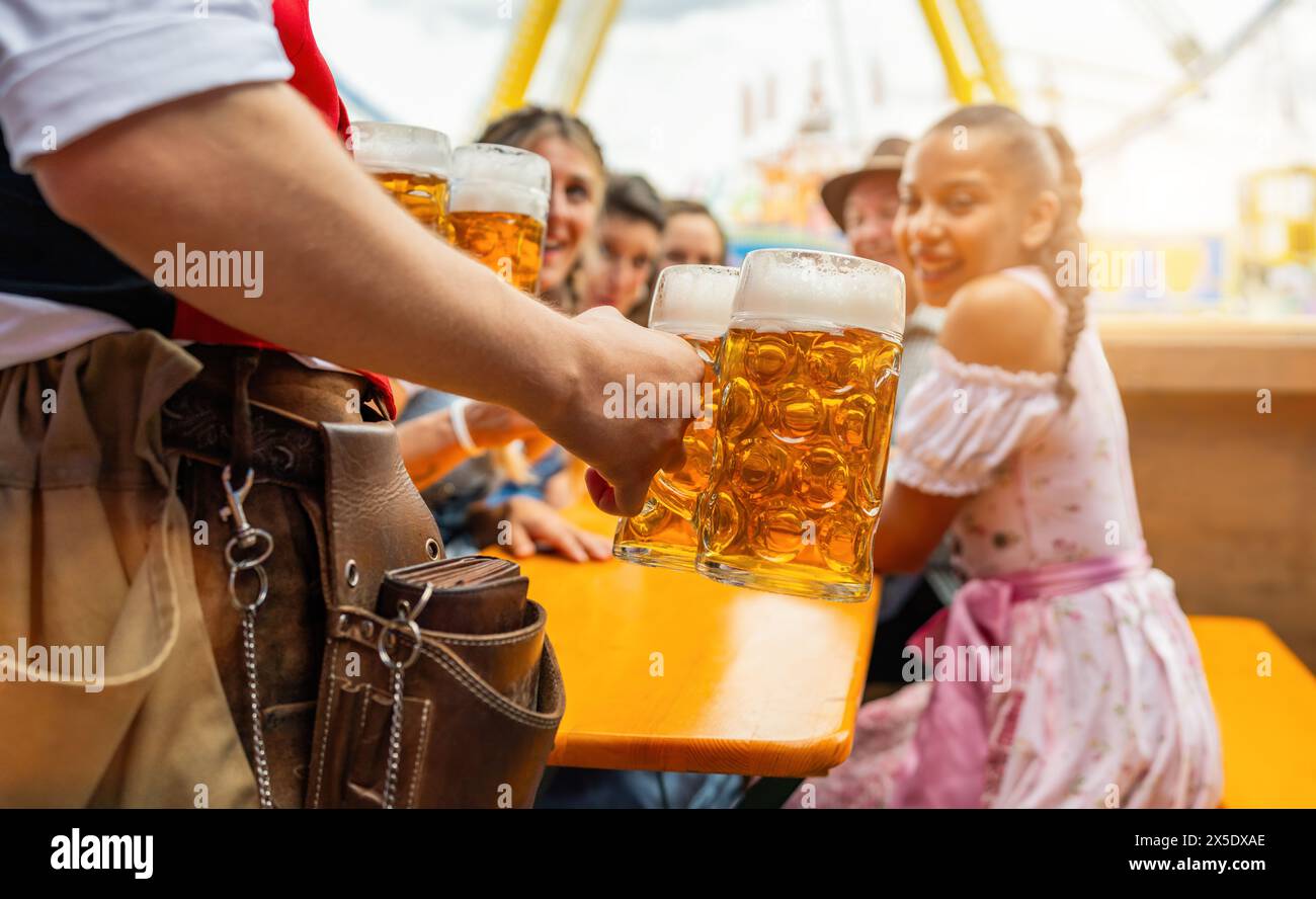 Cameriere che serve birra agli amici felici all'oktoberfest o al festival dult a un tavolo, giostre nel parco divertimenti sullo sfondo soleggiato Foto Stock