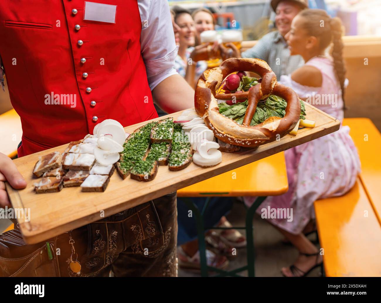 Cameriere in gilet rosso che serve piatti tradizionali tedeschi su un piatto di legno a un gruppo di persone in una tenda della birra a un festival dell'oktoberfest o dult in tedesco Foto Stock