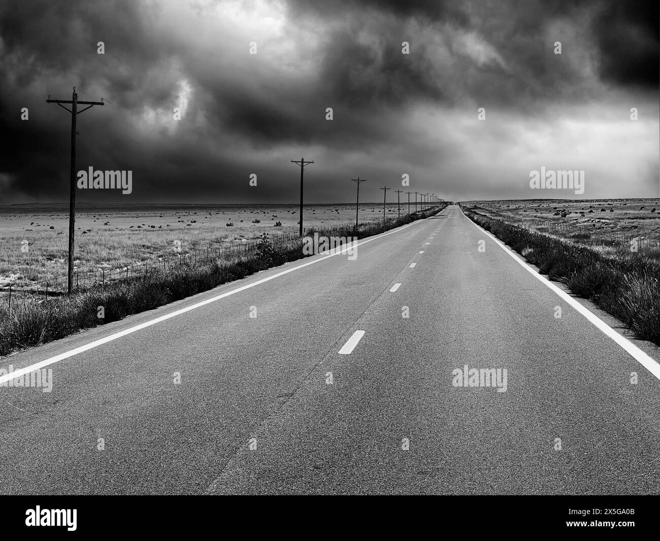 Un paesaggio bianco e nero che mostra una strada di campagna a due corsie vuota che si estende in lontananza con nuvole di tempesta in avvicinamento. Foto Stock