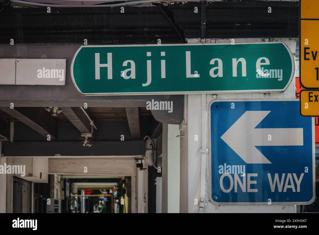 Un'immagine vivida che mostra un cartello stradale verde per Haji Lane accanto a una freccia direzionale blu a senso unico sotto un ponte urbano. Foto Stock