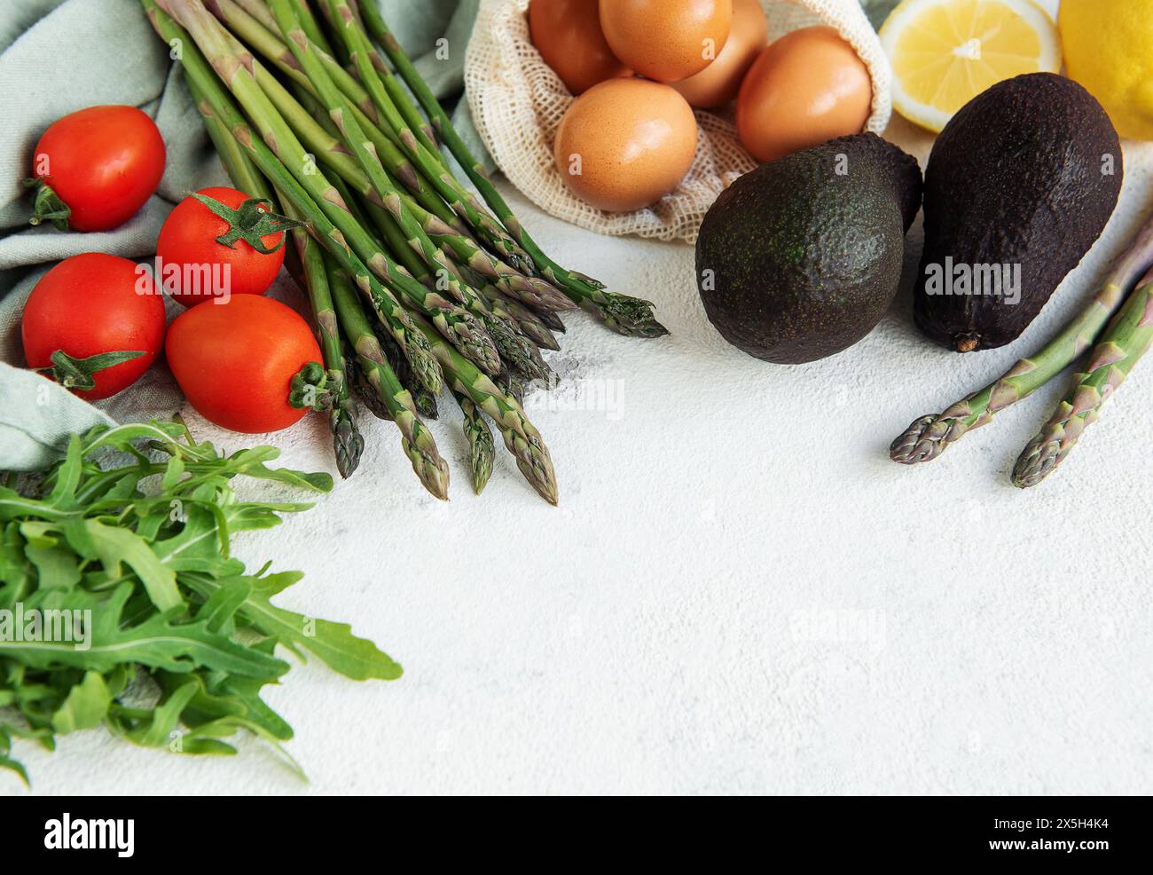 Una varietà di verdure fresche, tra cui asparagi, pomodori e rucola, sono ordinatamente organizzati accanto a avocado maturi e un sacchetto di uova scure. L'arr Foto Stock