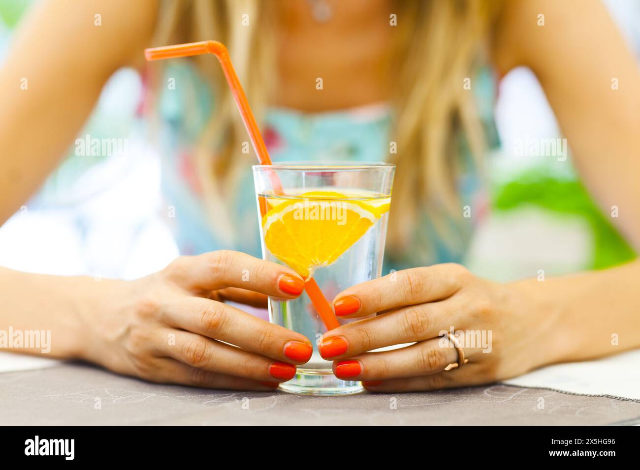 Primo piano delle mani di una donna che regge un bicchiere d'acqua guarnita con una fetta di arancia fresca, simboleggia uno stile di vita sano e l'idratazione. Foto Stock