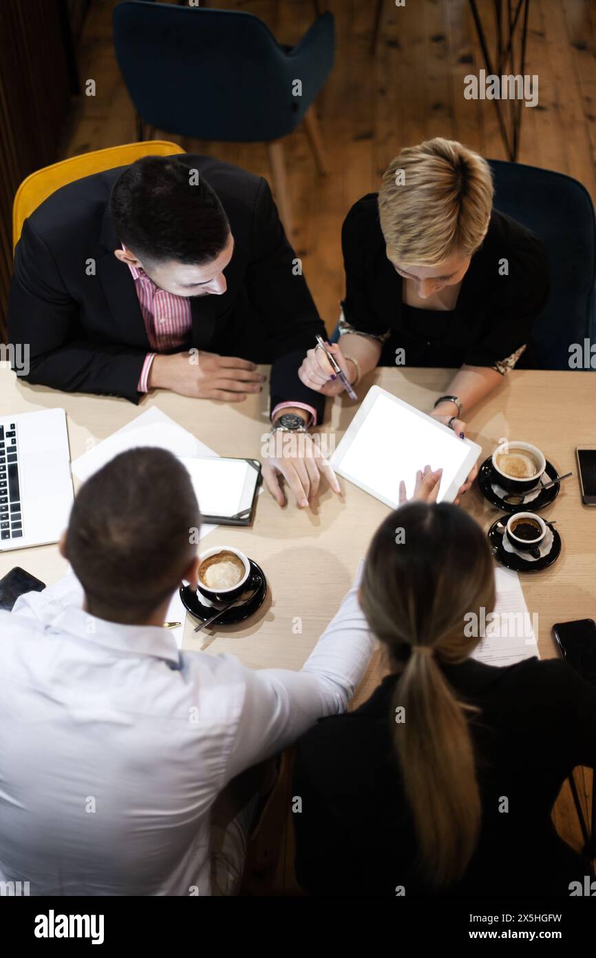 Ripresa aerea di una riunione professionale di lavoro in cui tre colleghi collaborano su documenti e dispositivi digitali in un ufficio moderno. Foto Stock