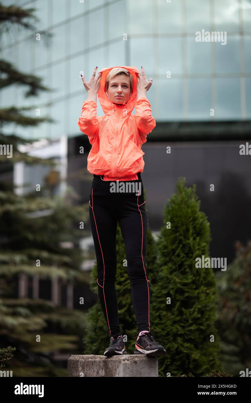 Una giovane donna sta su un piedistallo in città, regolando il cappuccio della sua giacca sportiva arancione brillante, trasmettendo un senso di fitness e stile urbano. Foto Stock