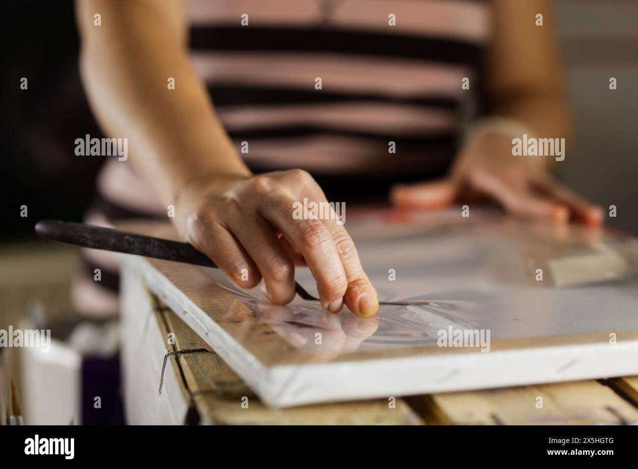 Vista ravvicinata delle mani di un'artista femminile mentre lavora con precisione su una tela, circondata da strumenti di pittura. Foto Stock