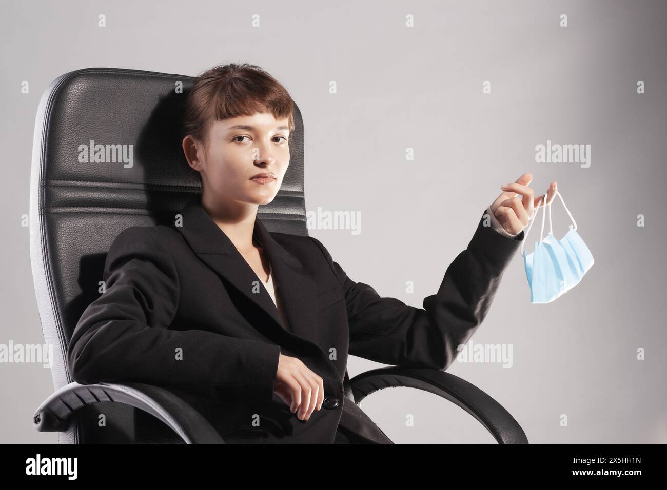 Giovane donna seria in abbigliamento da lavoro siede su una sedia da ufficio, tenendo una maschera medica blu, simboleggiando gli adattamenti sul posto di lavoro dovuti a problemi di salute. Foto Stock