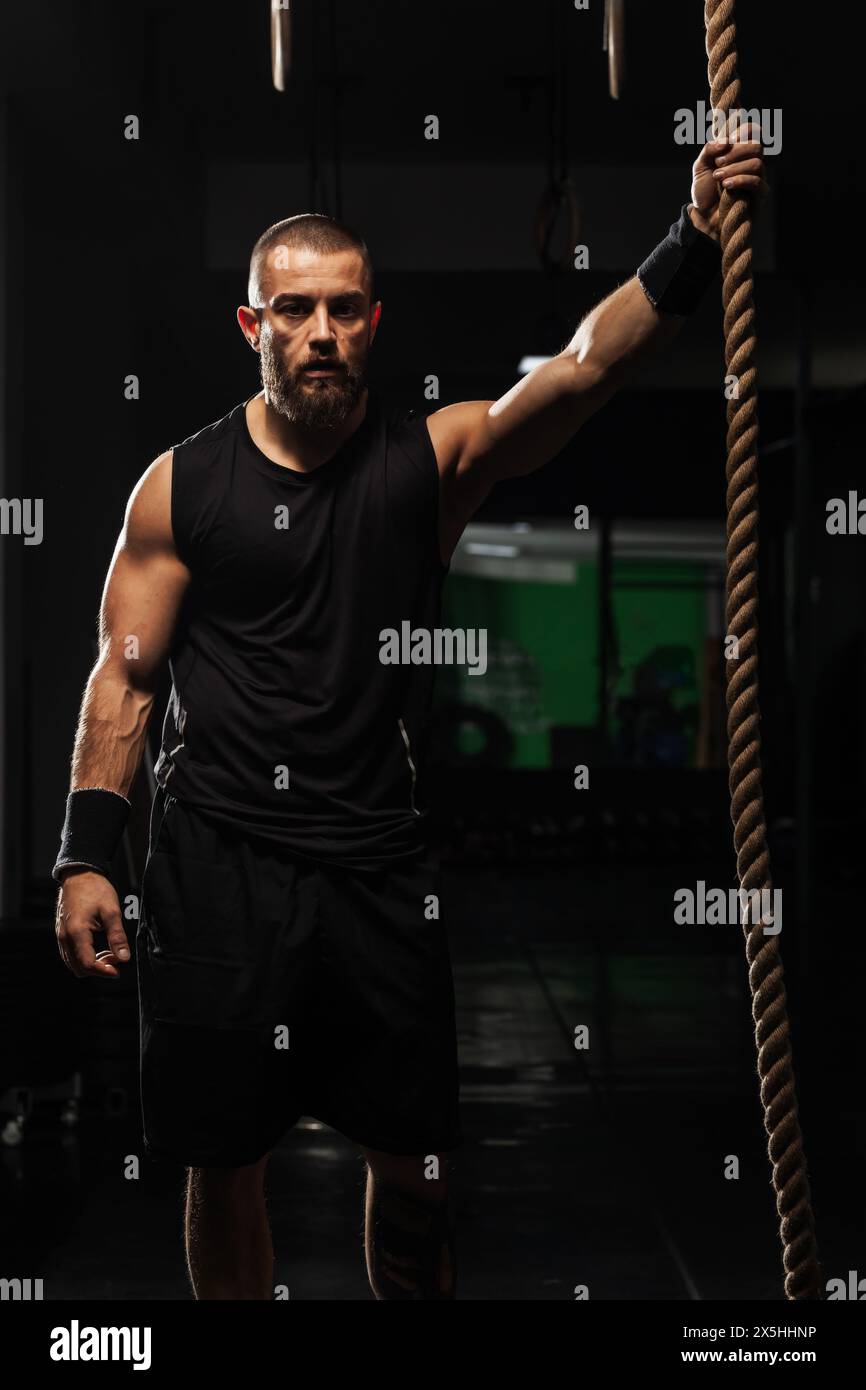 Un uomo muscoloso con la barba è pronto a salire su una corda in una palestra ben attrezzata, trasudando determinazione e forza. Foto Stock